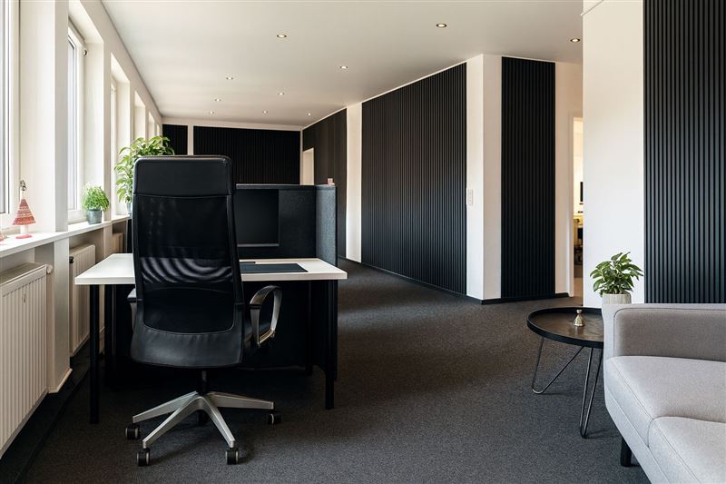 Heller Open Space mit Schreibtisch am Fenster, dunklen Lamellenwänden und Sitzecke