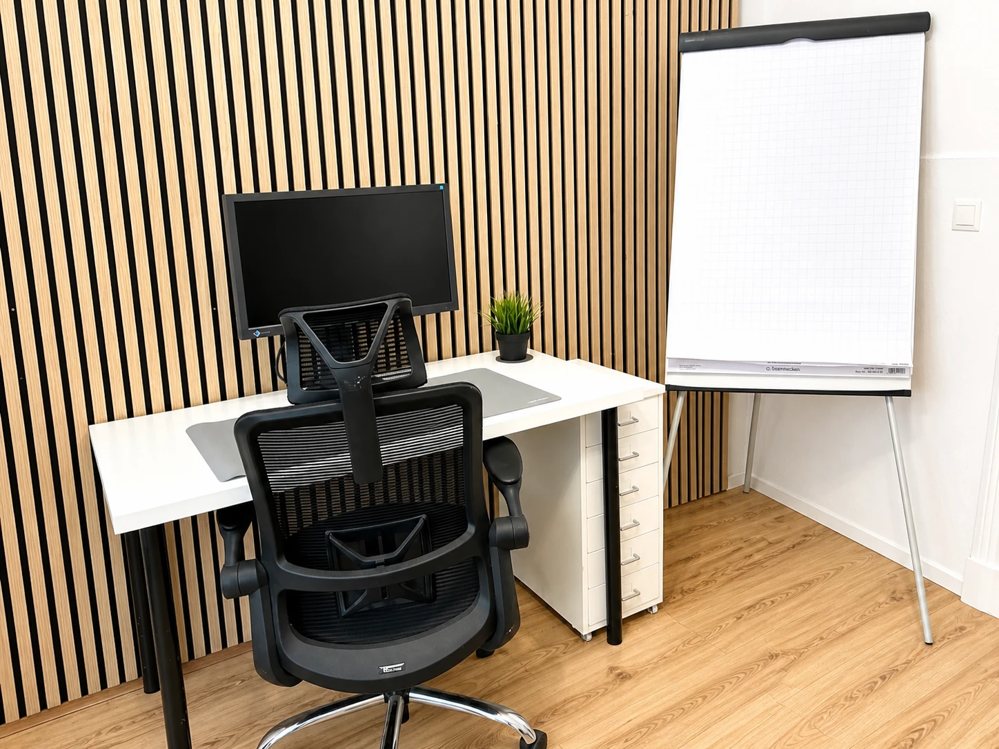 Arbeitsplatz am Standort Altona: weißer Schreibtisch mit Monitor, ergonomischer Bürostuhl und Flipchart vor einer Holzlamellenwand